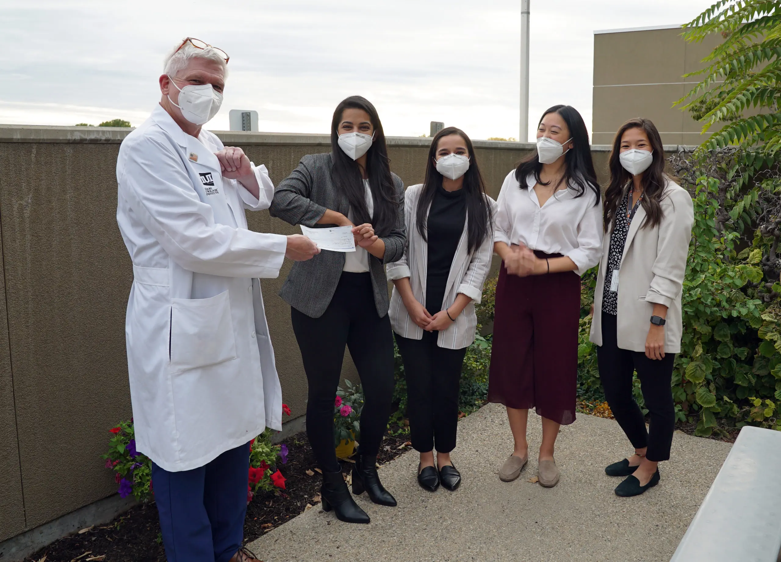 A doctor in a white coat and a mask hands a check to four women, also in masks. They are standing outside on a walkway with plants in the background, appearing happy and engaged in the moment.