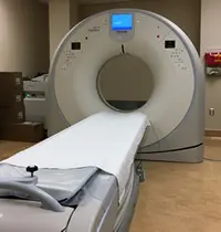 A medical CT scanner with a narrow bed positioned in front of a large circular opening is located in a hospital room. Cardboard boxes are stacked in the corner, and a wall-mounted fire extinguisher is visible in the background.